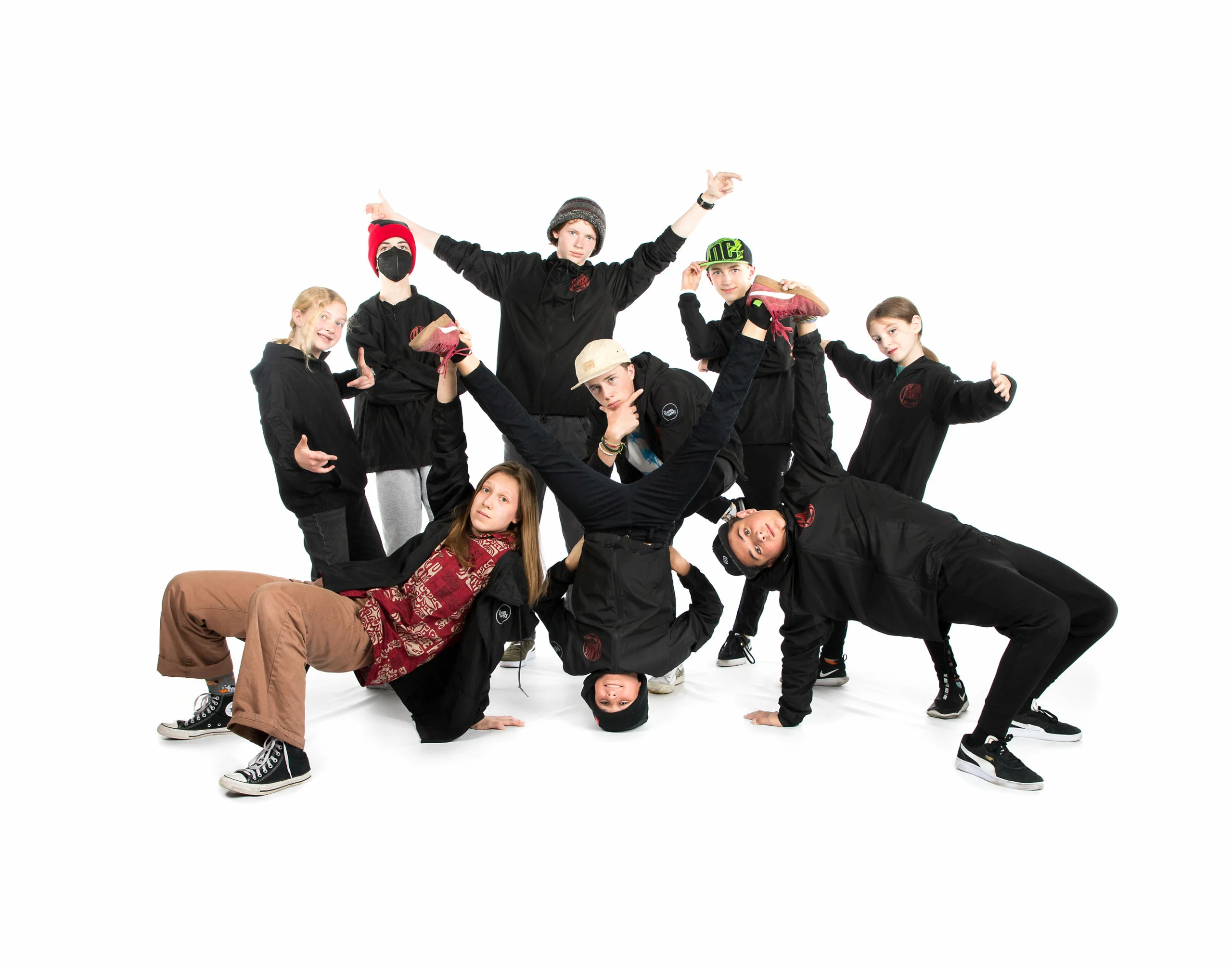 Nine dancers in black outfits pose energetically against a plain white background.