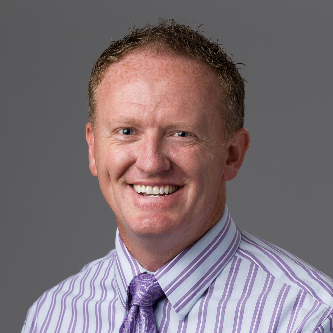 Smiling man with short red hair in a striped shirt and purple tie on a gray background.