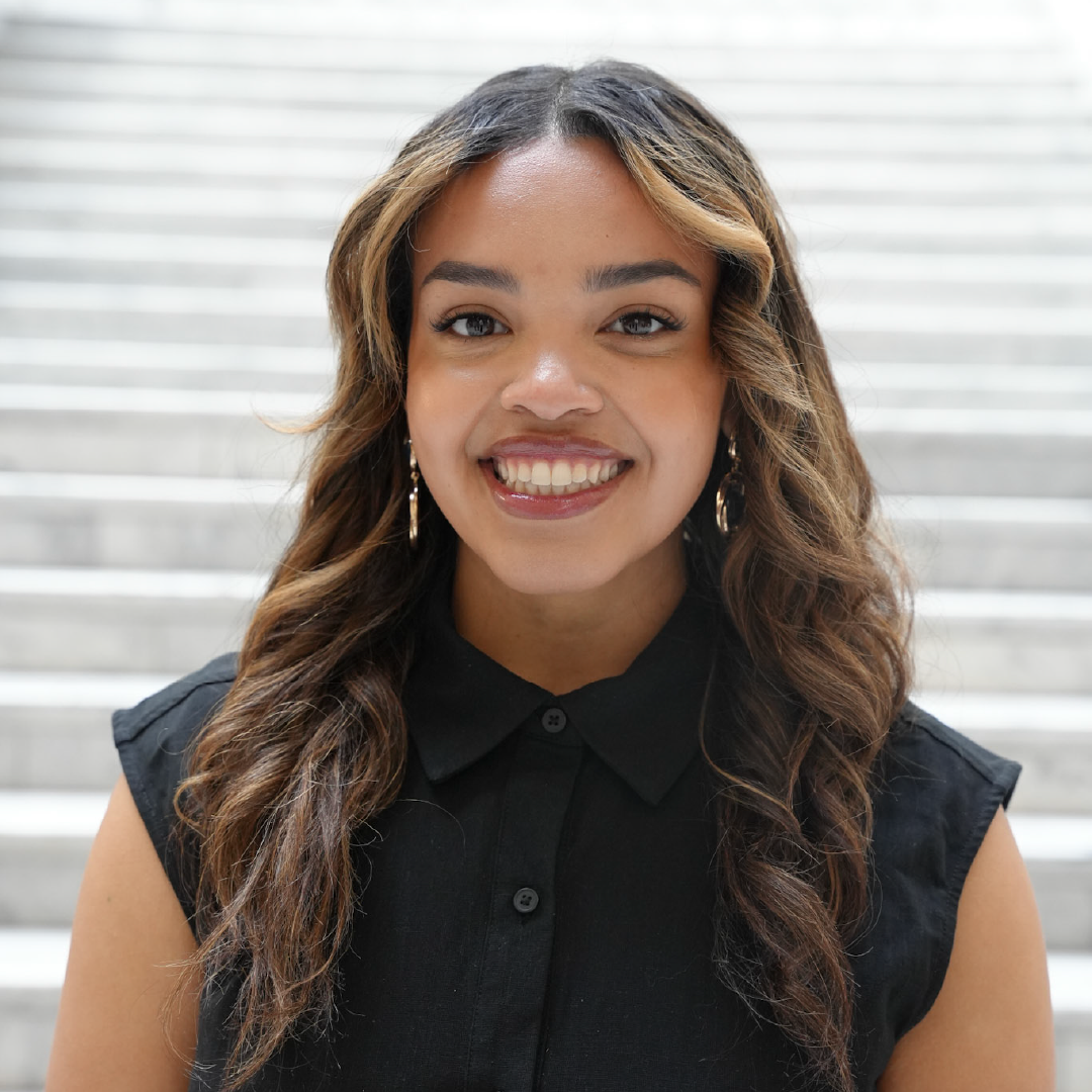 Smiling woman with wavy hair in a black shirt standing in front of marble stairs.