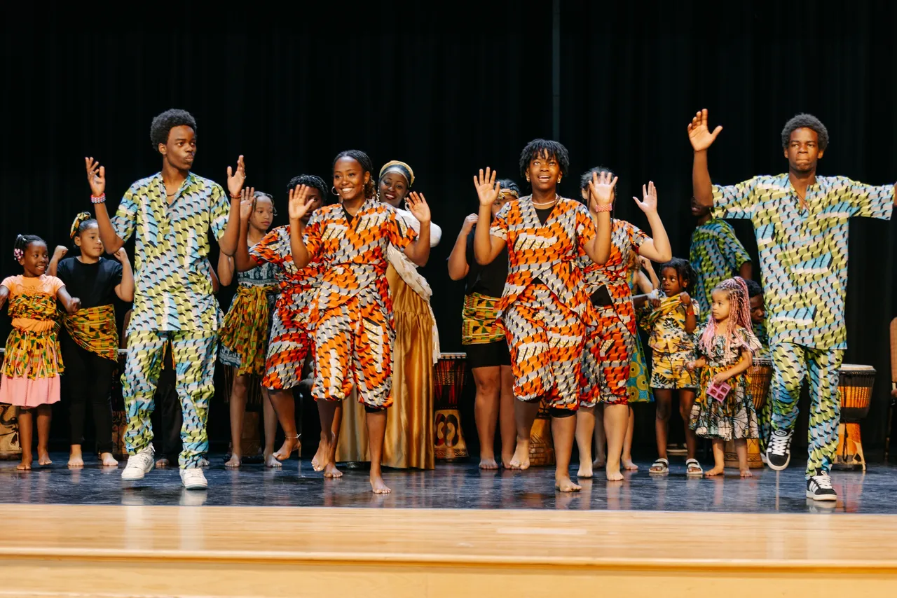 A group of people in colorful attire perform a dance on stage with hands raised.