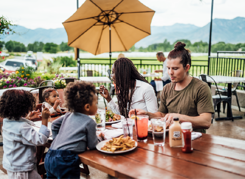 Family eating together at an outdoor restaurant table with food and drinks under an umbrella.