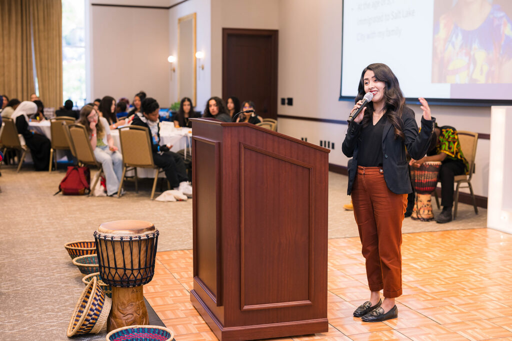 A woman speaks at a podium in a conference room with tables and African drums nearby.