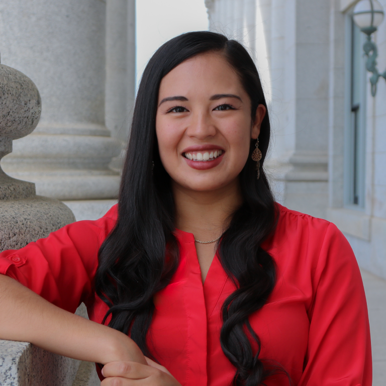 Woman with long dark hair in a red blouse smiling, standing outside near a stone building.