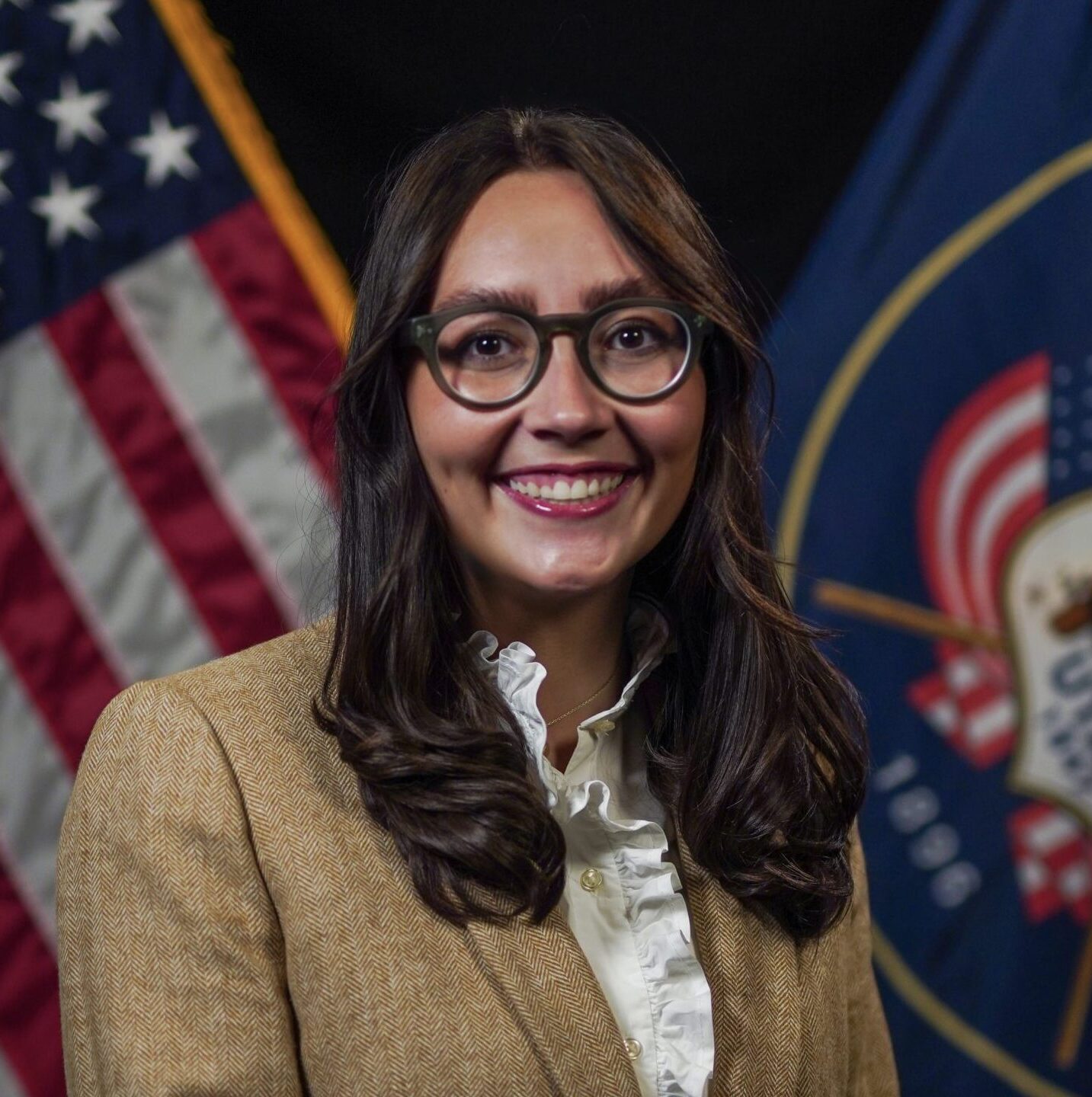 Woman in tan blazer poses in front of American and Utah state flags against a dark background.